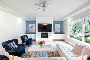 A modern living room with a fireplace, a ceiling fan, and large windows offering a view of the outdoors.