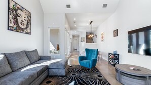 Uma sala de estar moderna com um sofá seccional cinza, uma poltrona azul, um tapete com padrão preto e branco e uma televisão grande na parede.