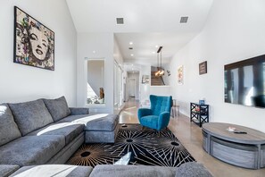 A modern living room with a grey sectional sofa, a blue armchair, a black and white patterned rug, and a large TV on the wall.
