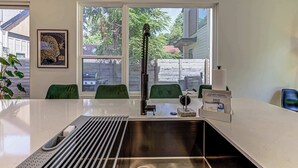 A kitchen with a stainless steel sink, green chairs, a window with a view of a house, and a sign offering grab-and-go goodies.