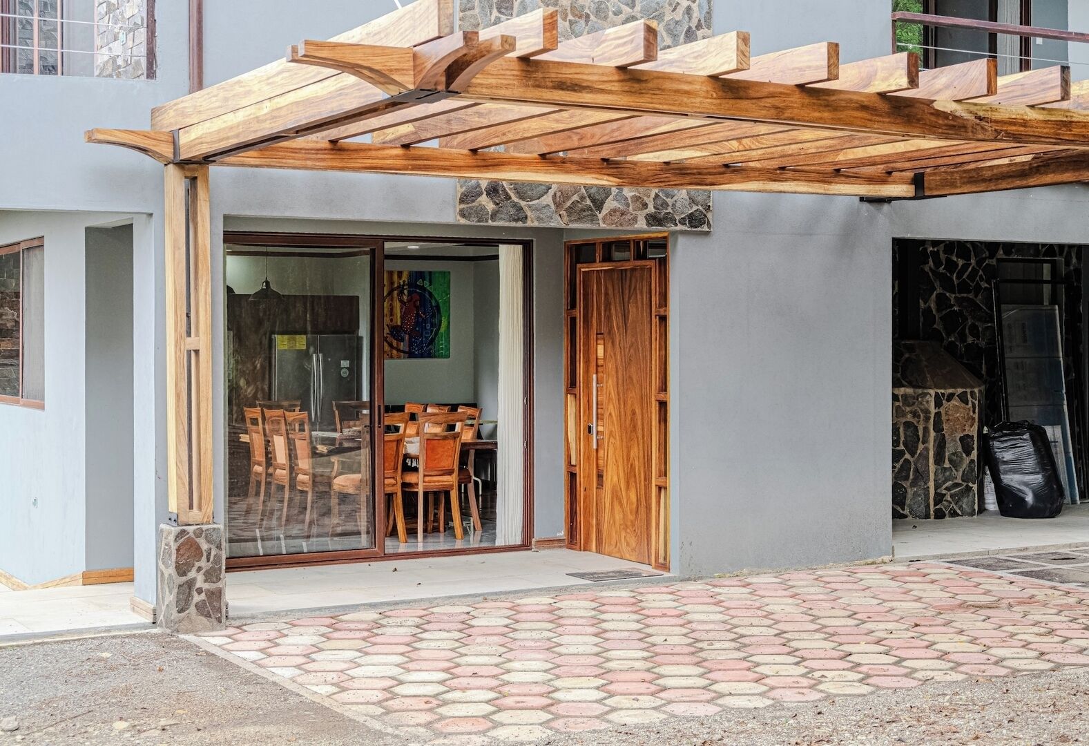 Une entrée d’immeuble agrémentée d’une pergola en bois, une porte vitrée donnant sur un espace intérieur comprenant un coin repas, et un mur en pierre.