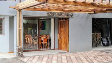 Une entrée d’immeuble agrémentée d’une pergola en bois, une porte vitrée donnant sur un espace intérieur comprenant un coin repas, et un mur en pierre.