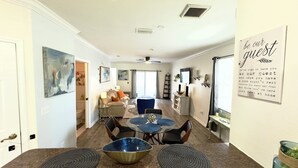 A spacious living area with a dining table, chairs, and a sofa. There are paintings on the walls, a ceiling fan, and a window with curtains.