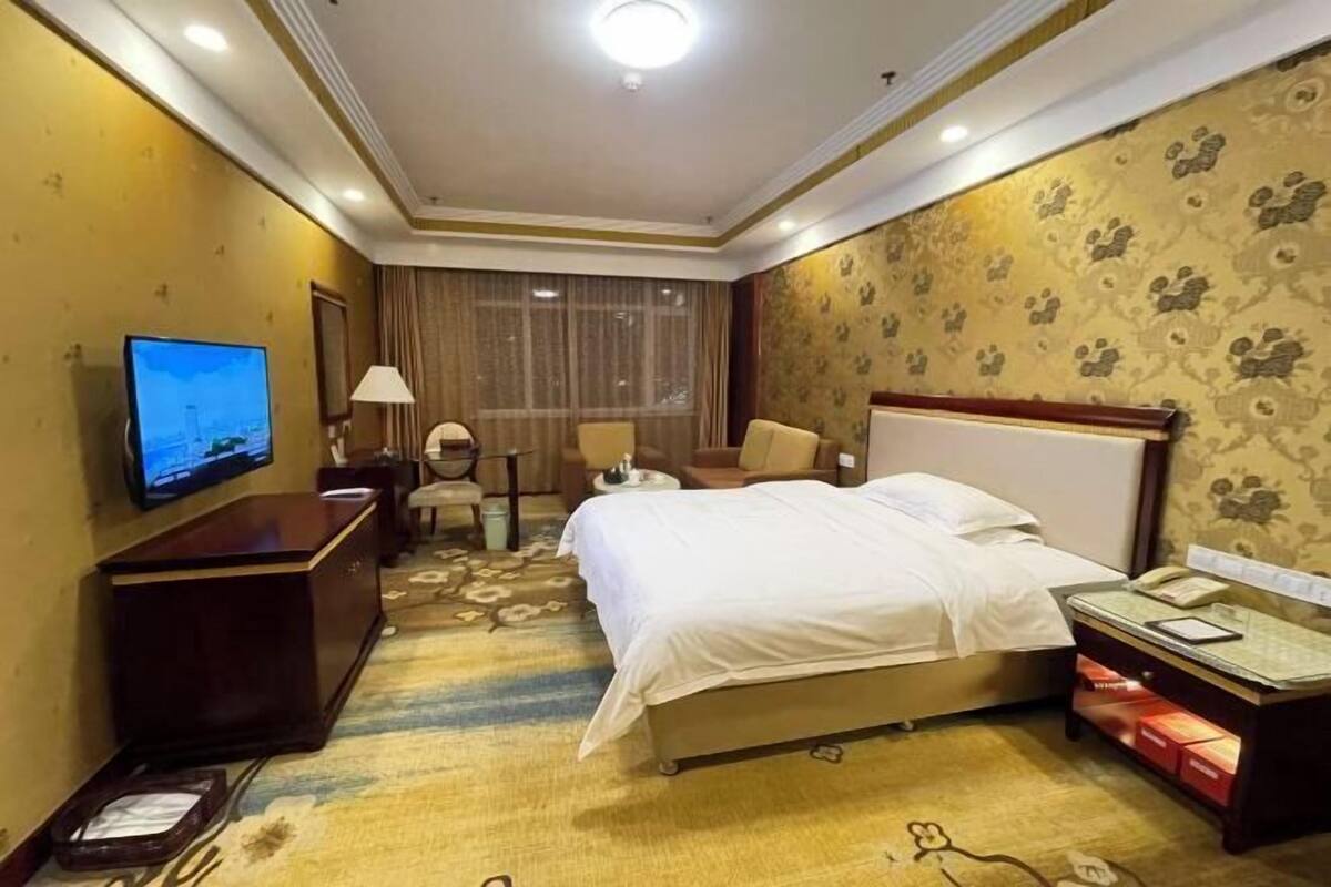 A hotel room with a bed, a desk with a television, a chair, a lamp, and a nightstand with a phone and a lamp.
