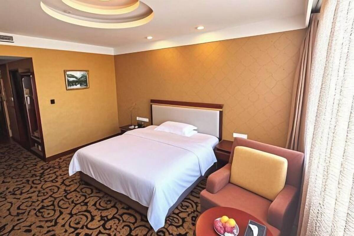 A hotel room with a bed, a nightstand, a chair, and a small table with fruit.
