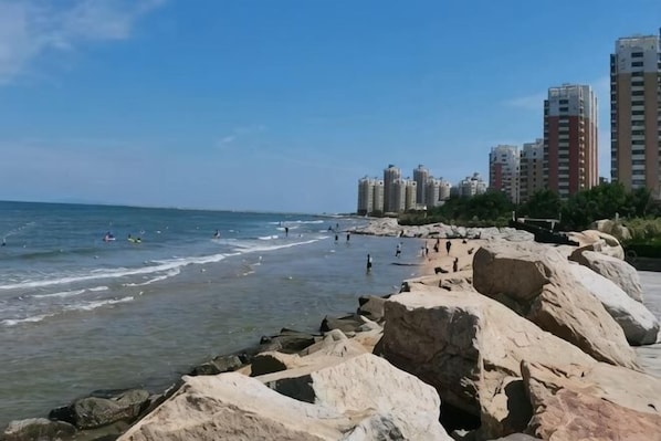 A beach with rocky shoreline, clear blue sky, and high-rise buildings in the background. - Gulf Hotel (Longkou)