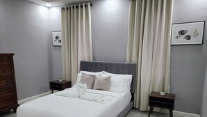 A neatly made bed with white linens and decorative swan pillows, a grey upholstered headboard, and a small bedside table with a plant. - Modern Sunset Oasis - 2BD Apt w/ Pool & Views, 5 Min to Dining & Shopping (Kingston)