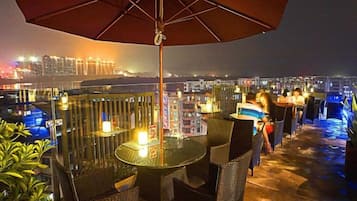 An outdoor terrace with tables and chairs, a large umbrella, and a cityscape view in the background.