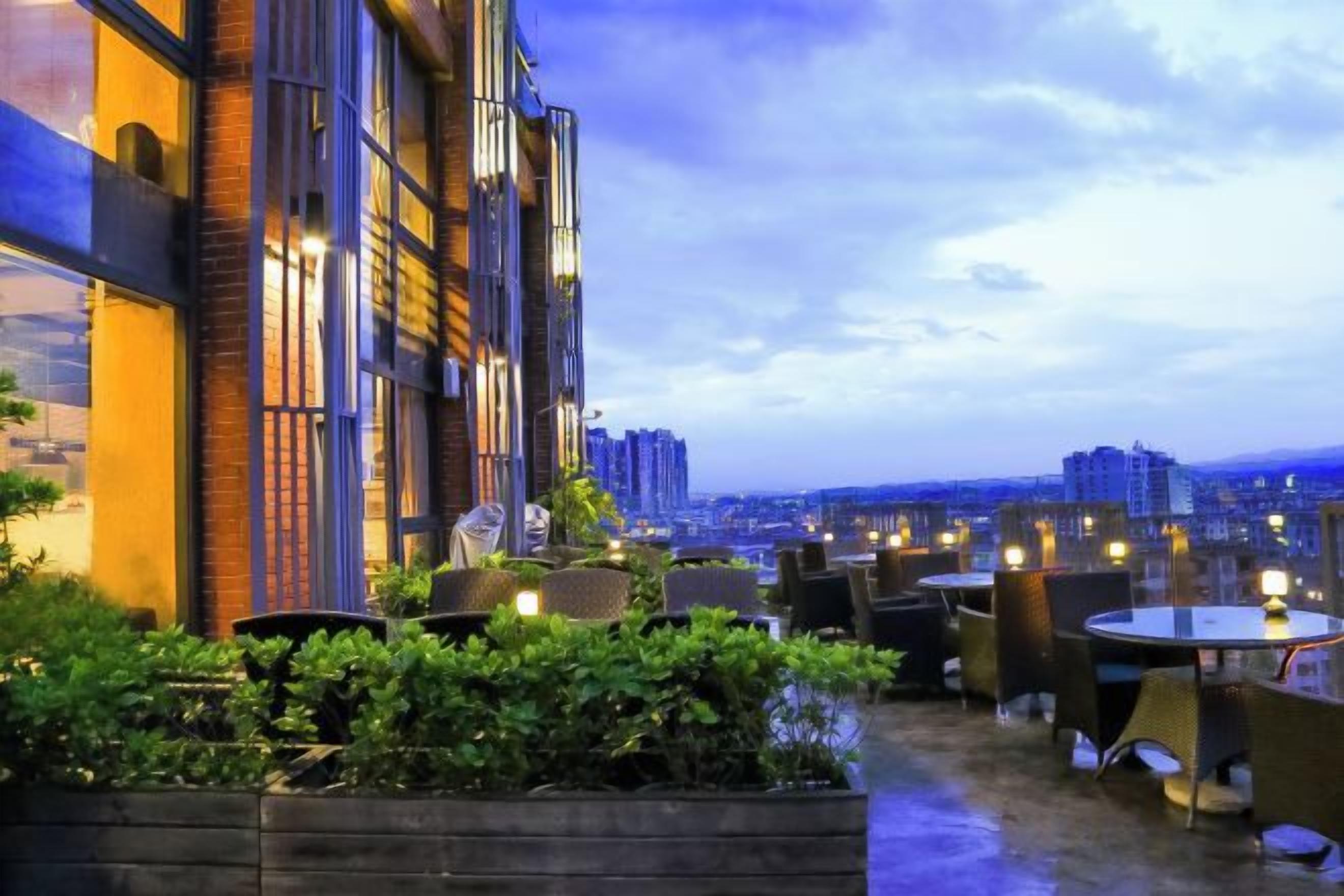 Uma área de estar ao ar livre com mesas e cadeiras, cercada por vegetação, com vista para uma paisagem urbana.