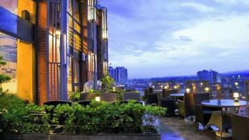 An outdoor seating area with tables and chairs, surrounded by greenery, and overlooking a cityscape.