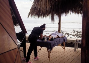 A wooden deck with a massage table, a person attending to a client, and a beach view in the background.