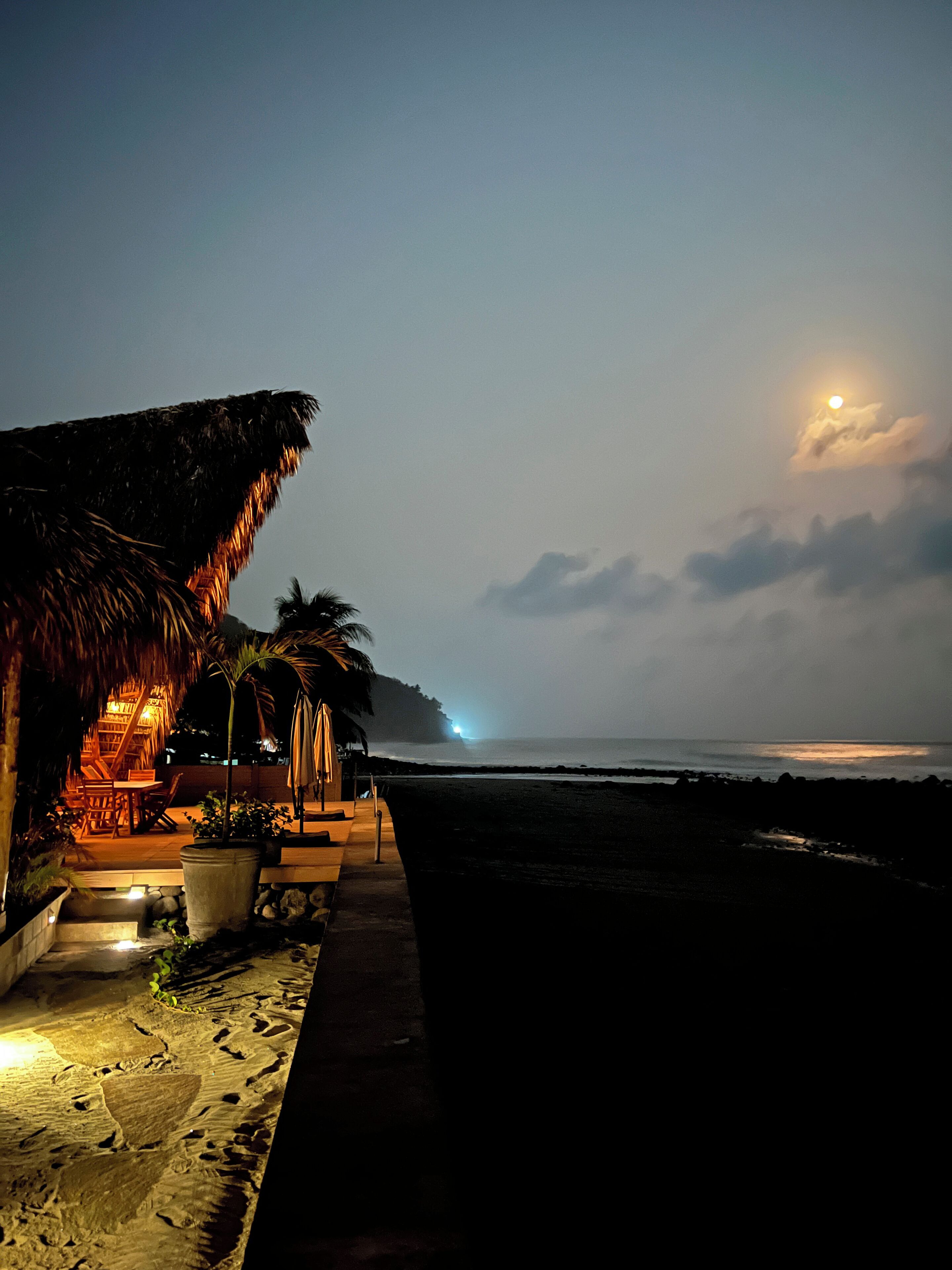 A beachside hut with a thatched roof, a small outdoor seating area, and a view of the ocean at night.