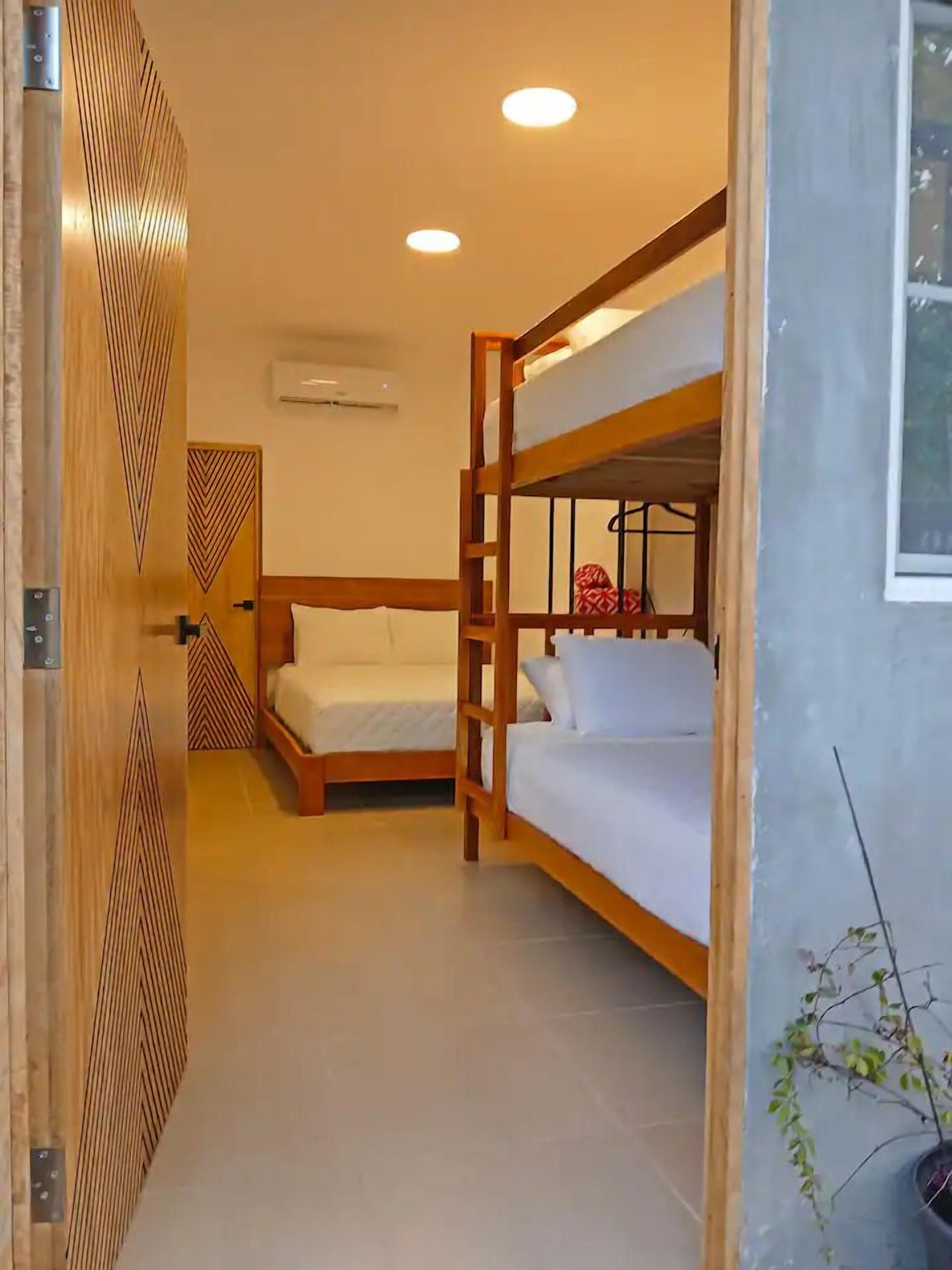 Quarto com duas beliches, uma porta de madeira com padrão geométrico, uma pequena planta em vaso e uma janela.