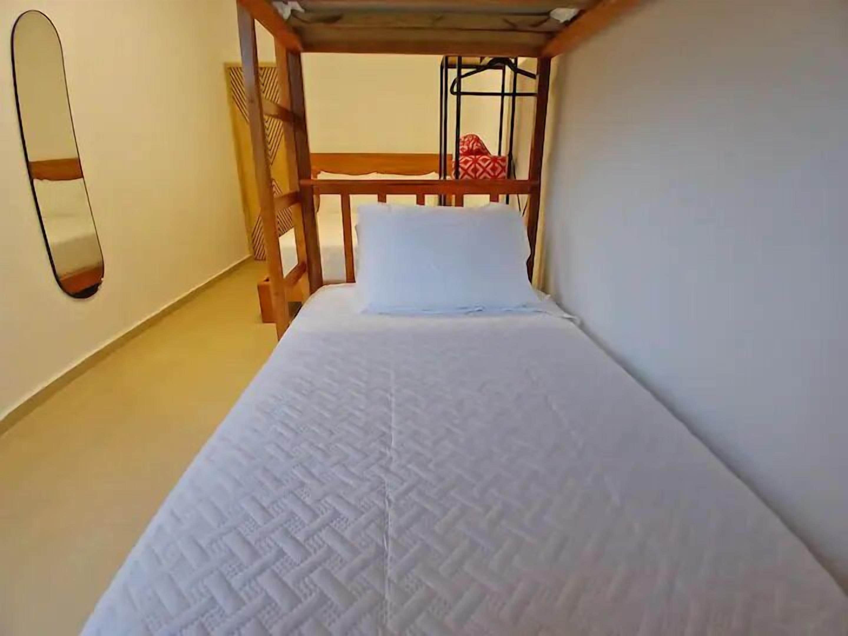A narrow room with a single bed, a wooden headboard, a mirror on the wall, and a small red patterned item on the headboard.