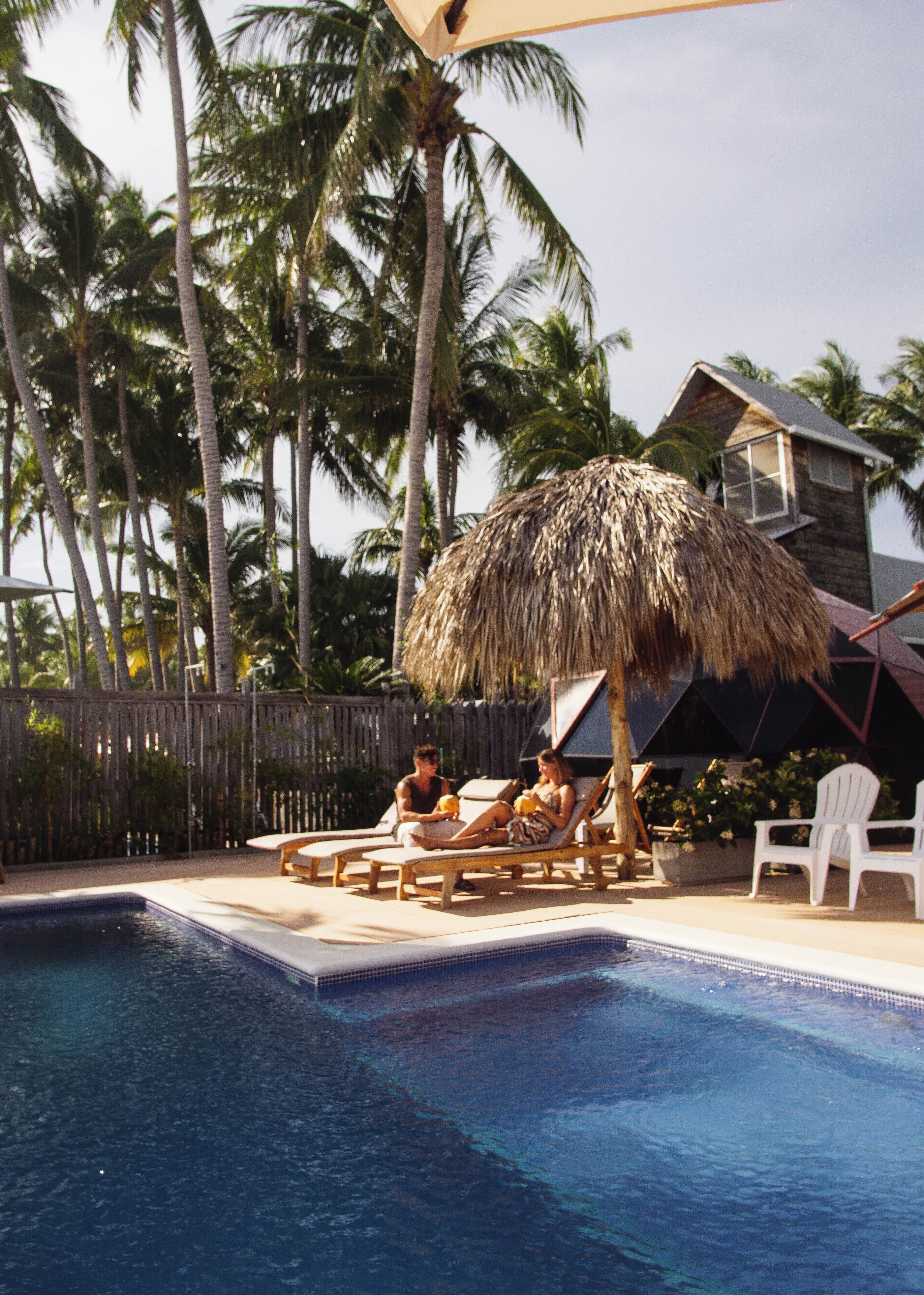 A pool area with lounge chairs and a thatched umbrella, surrounded by palm trees and a wooden fence.