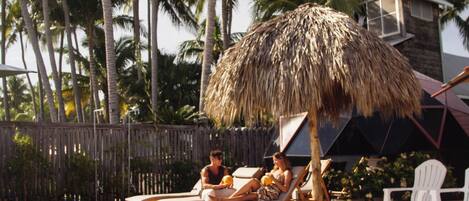 A pool area with lounge chairs and a thatched umbrella, surrounded by palm trees and a wooden fence.