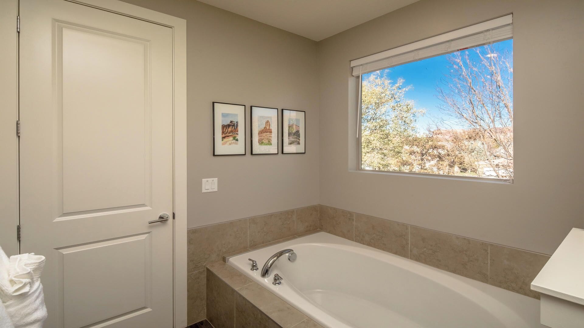 A bathroom with a bathtub, a window with a view of trees, and three framed pictures on the wall.