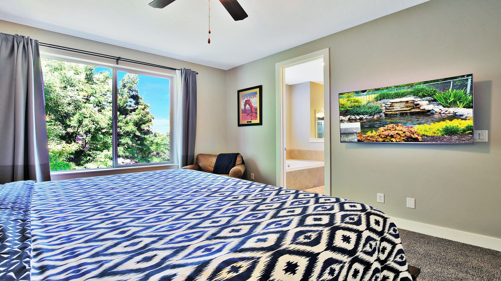 A bedroom with a large bed, a ceiling fan, a TV on the wall, and a view of trees through the window.