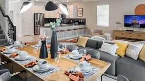 Uma sala de jantar moderna com uma mesa de madeira posta para refeição, uma cozinha ao fundo e uma sala de estar com sofá e televisão.