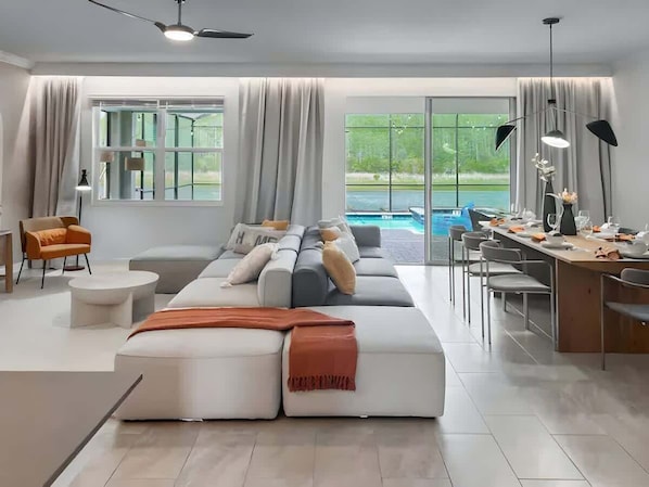 A modern living room with a large sofa, a dining table, and a view of a swimming pool.