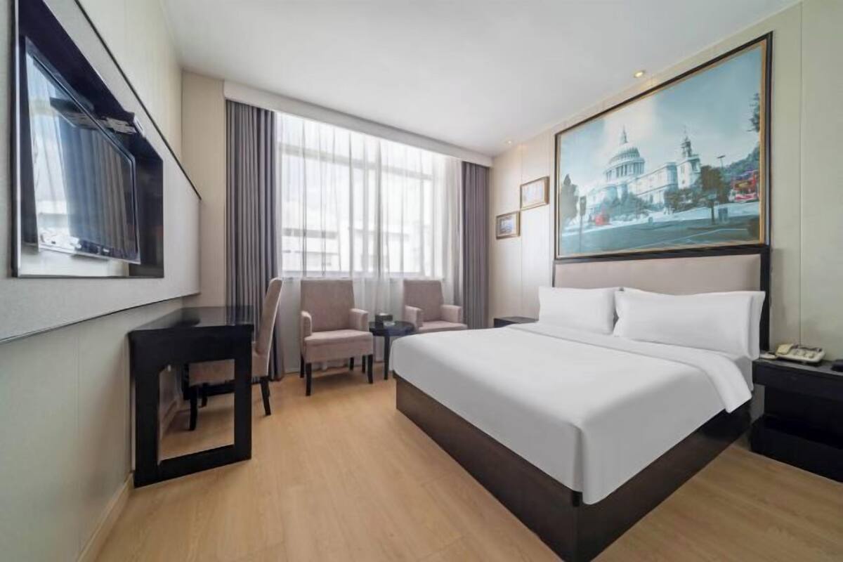 A hotel room with a large bed, two armchairs, a small table, a TV, and a painting of a building.