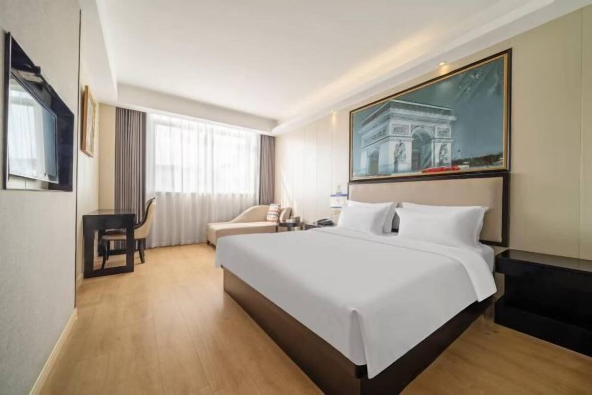 A hotel room with a large bed, a desk, a chair, a window with curtains, and a painting of the Arc de Triomphe.