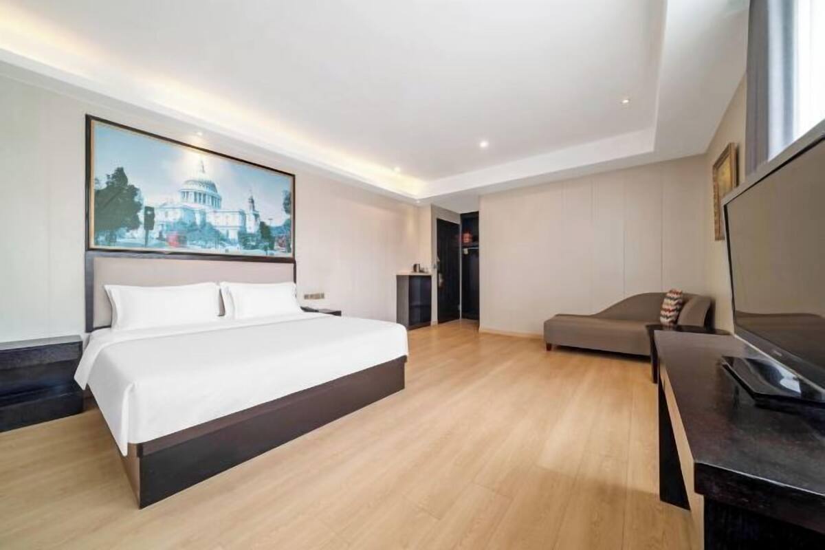 A modern hotel room with a large bed, a sofa, a flat-screen TV, and a painting of a building on the wall.