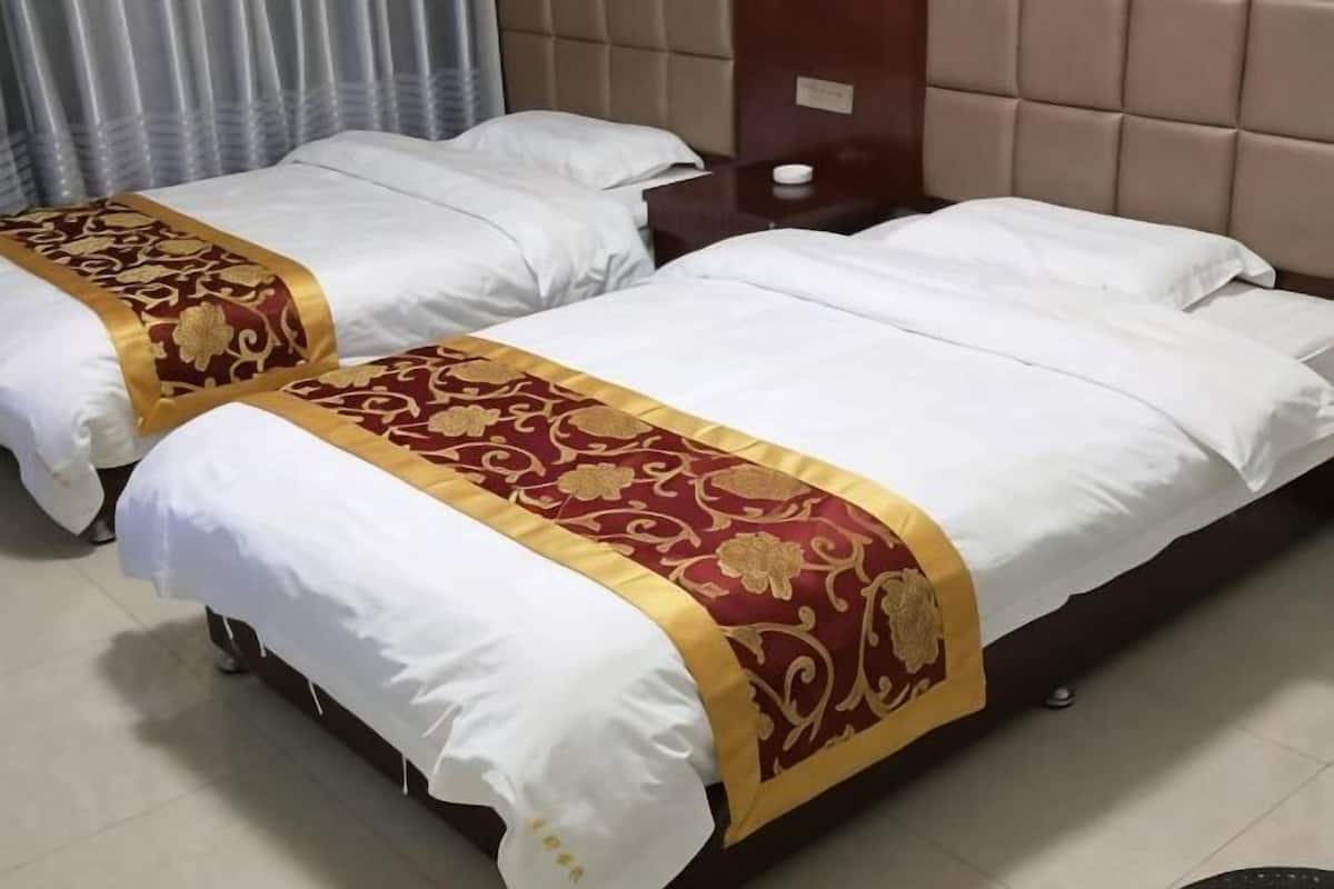 Two beds with white linens and red and gold bedspreads, a wooden headboard, and a small bedside table with a lamp.