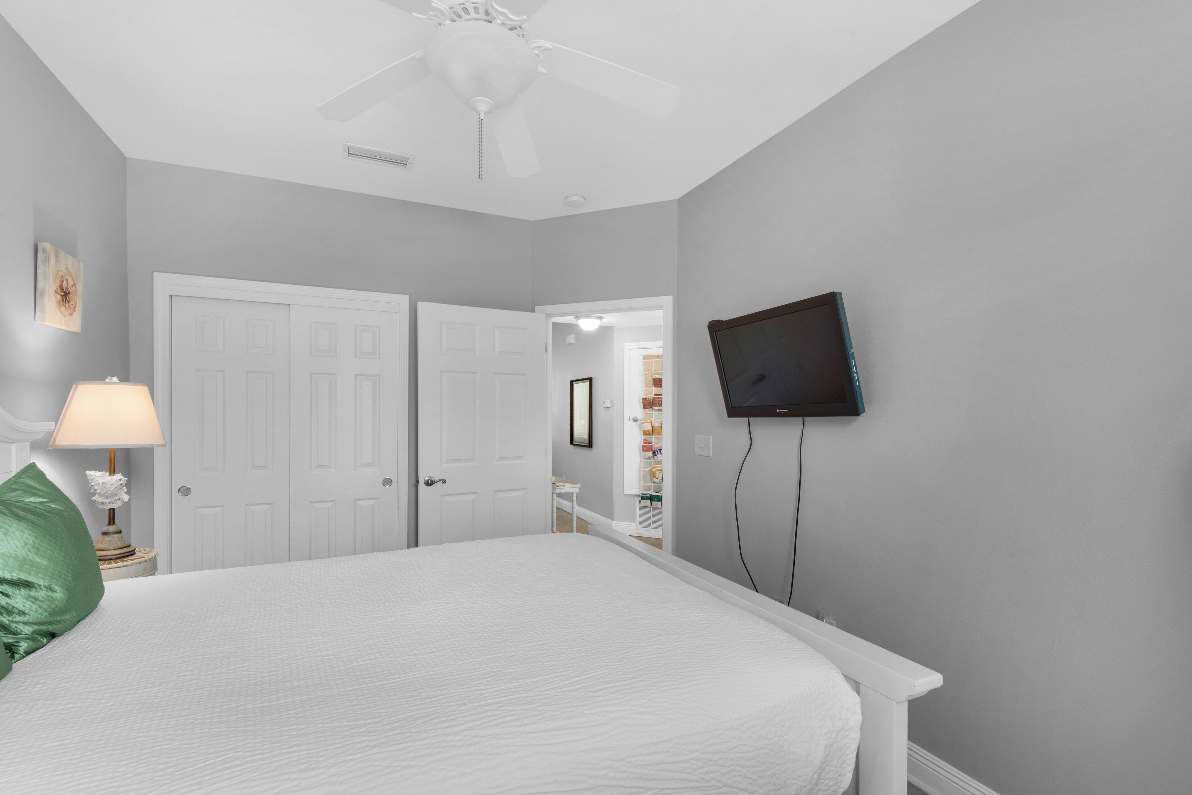 Ein Schlafzimmer mit Bett, Deckenventilator, an der Wand montiertem Fernseher und einer Tür zu einem anderen Raum.