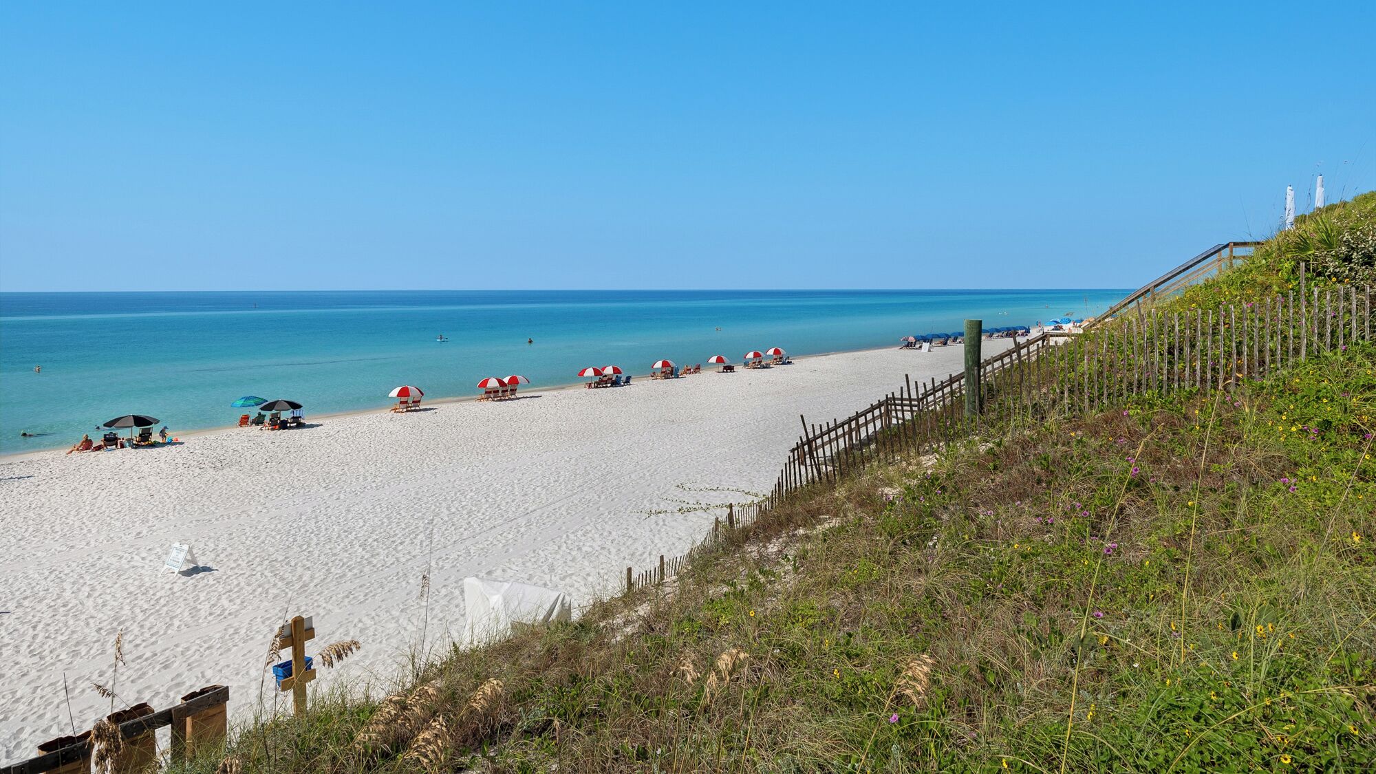 A beach with white sand, red and white umbrellas, and a wooden fence.