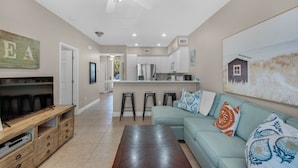 A modern living room with a sectional sofa, a wooden entertainment unit, and a dining area with bar stools. - The Village at Blue Mountain Beach (Santa Rosa Beach)
