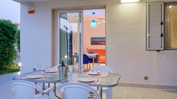 An outdoor dining area with a glass table and white chairs, a pergola overhead, and a view into the house. - Eolo, holiday home with garden 100 mt from the sea (Marina di Ragusa)