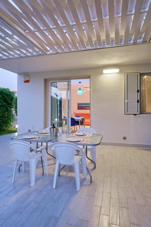 Een terras met een glazen tafel en witte stoelen, een pergola erboven en uitzicht op het huis.