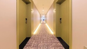 Een hotelgang met patroonbelegd tapijt, lichte muren en wandlampen.