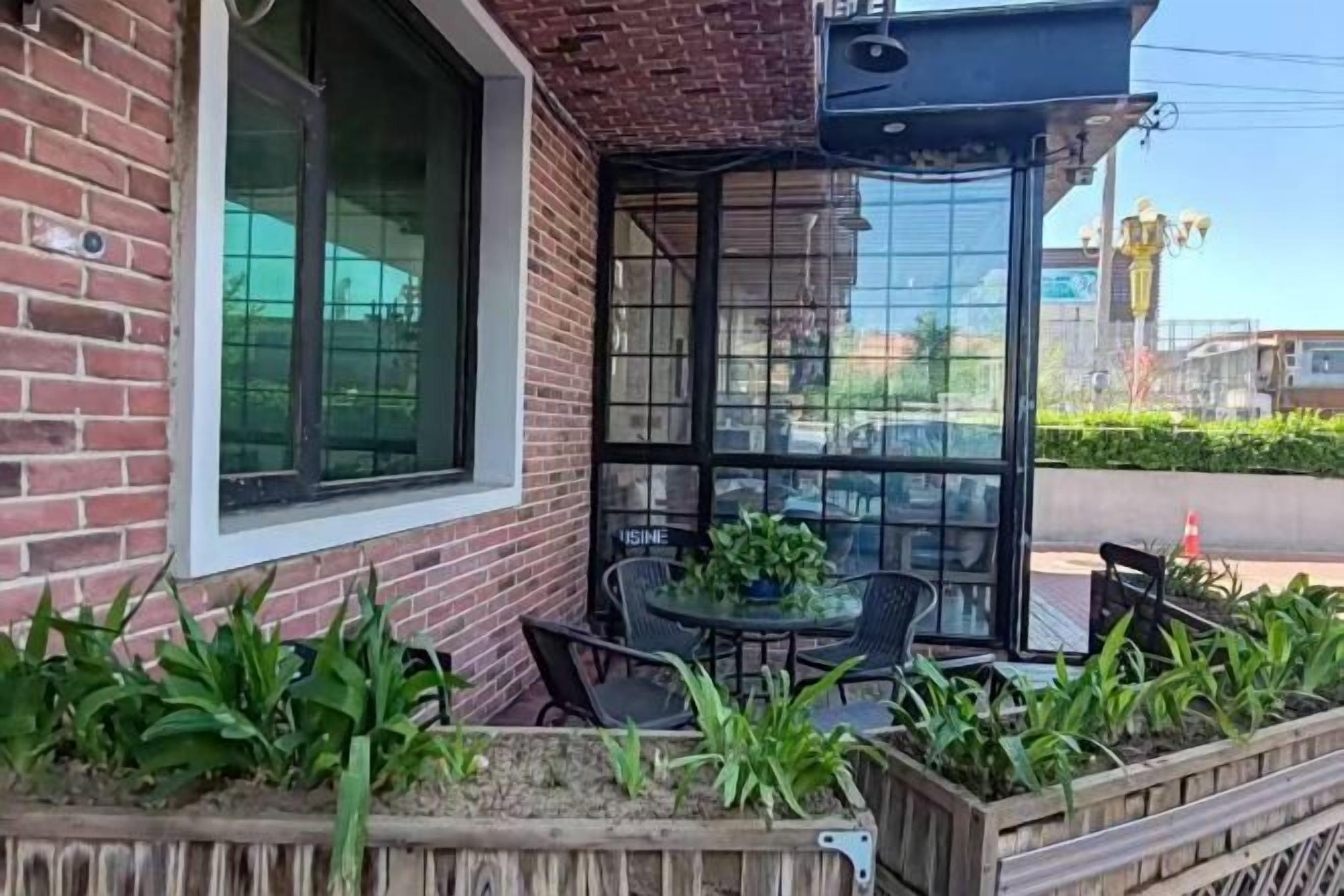 A brick building with a glass door entrance, outdoor seating with chairs and a table, and a planter with green plants.