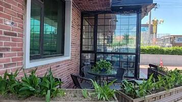 Un edificio de ladrillo con entrada por una puerta de cristal, mobiliario exterior con sillas y una mesa, y una jardinera con plantas verdes.