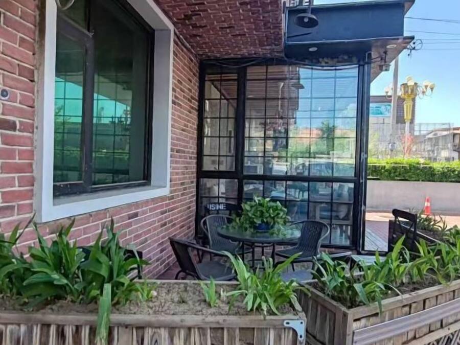 A brick building with a glass door entrance, outdoor seating with chairs and a table, and a planter with green plants.