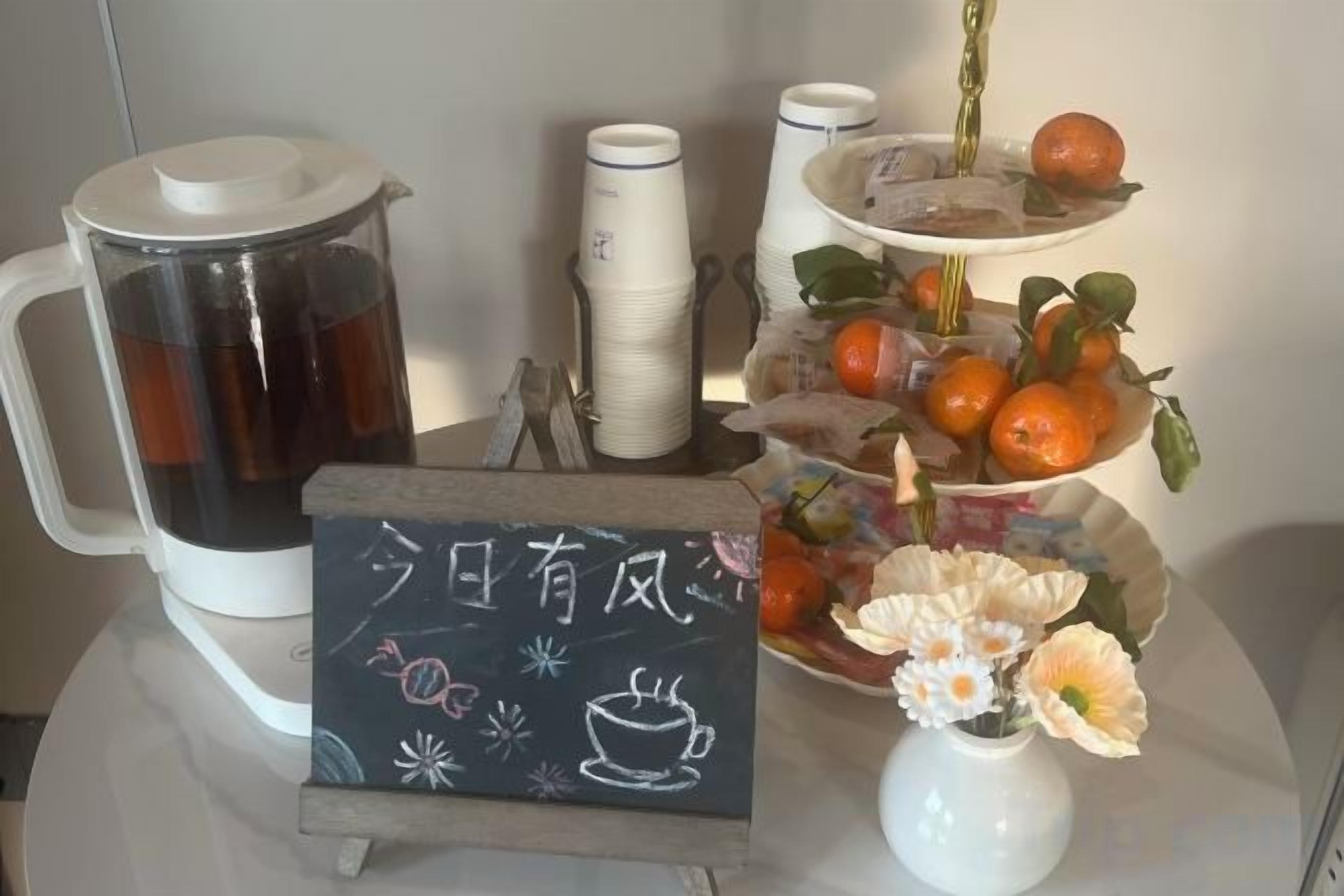 Una estación de café con cafetera, tazas y un frutero escalonado con naranjas y flores.