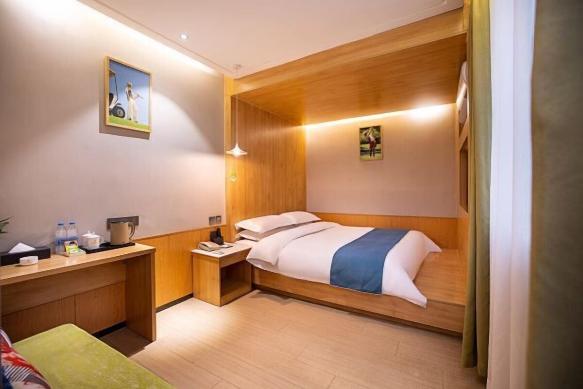 Ein Hotelzimmer mit Holz Kopfteil, einem Einzelbett, einem Nachttisch mit Lampe, einem kleinen Schreibtisch mit Stuhl und einer grünen Bank.