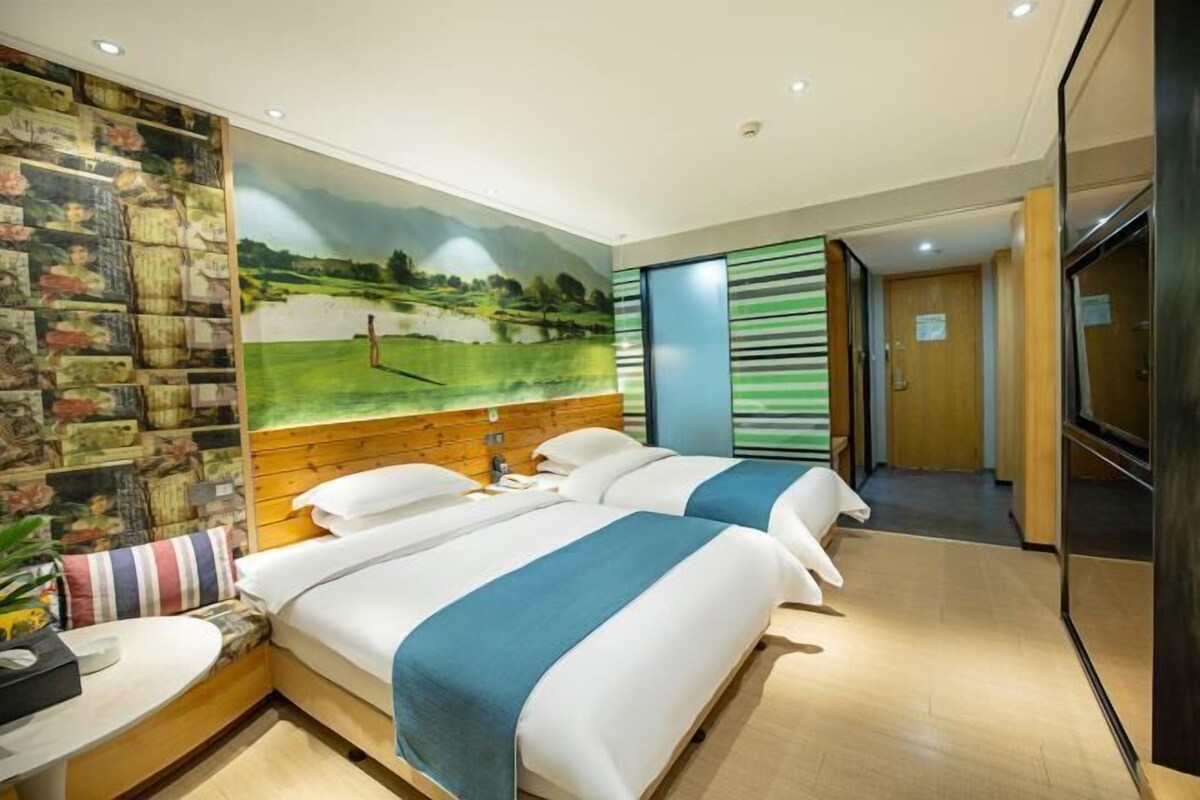 Ein Hotelzimmer mit zwei Betten, einem Wandbild, einem hölzernen Kopfteil, einem kleinen Tisch und einer Pflanze.