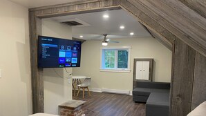 A modern room with a large flat-screen TV mounted on a wooden wall, a ceiling fan, a small table with a chair, a grey sofa, and a window with a view of trees. - Beautiful Waterfront Cottage with Hot Tub in South Muskoka  (Severn Bridge)