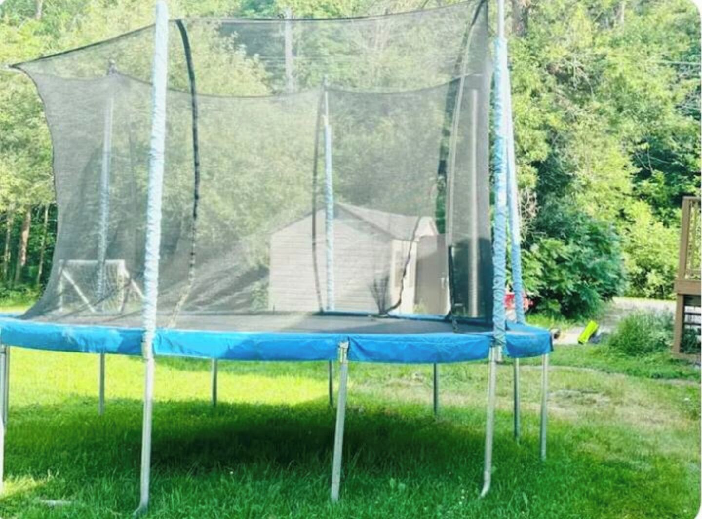 A trampoline with a safety net enclosure in a grassy area with trees in the background.