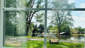 A view through a window of a grassy area with a lawn, a bench, and a small building near a body of water. - Beautiful Waterfront Cottage with Hot Tub in South Muskoka  (Severn Bridge)