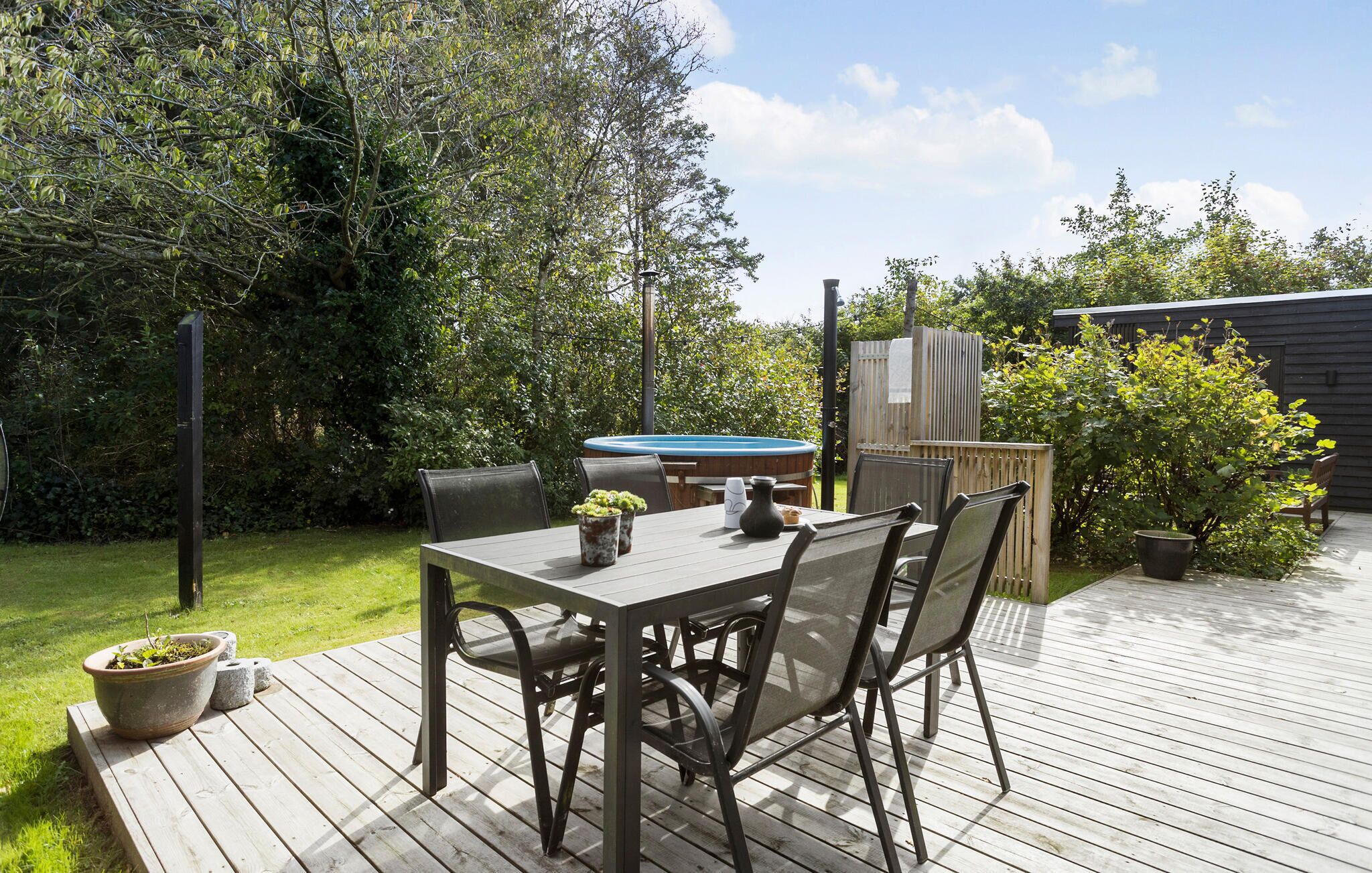 A wooden deck with a table and chairs, surrounded by greenery and a hot tub.