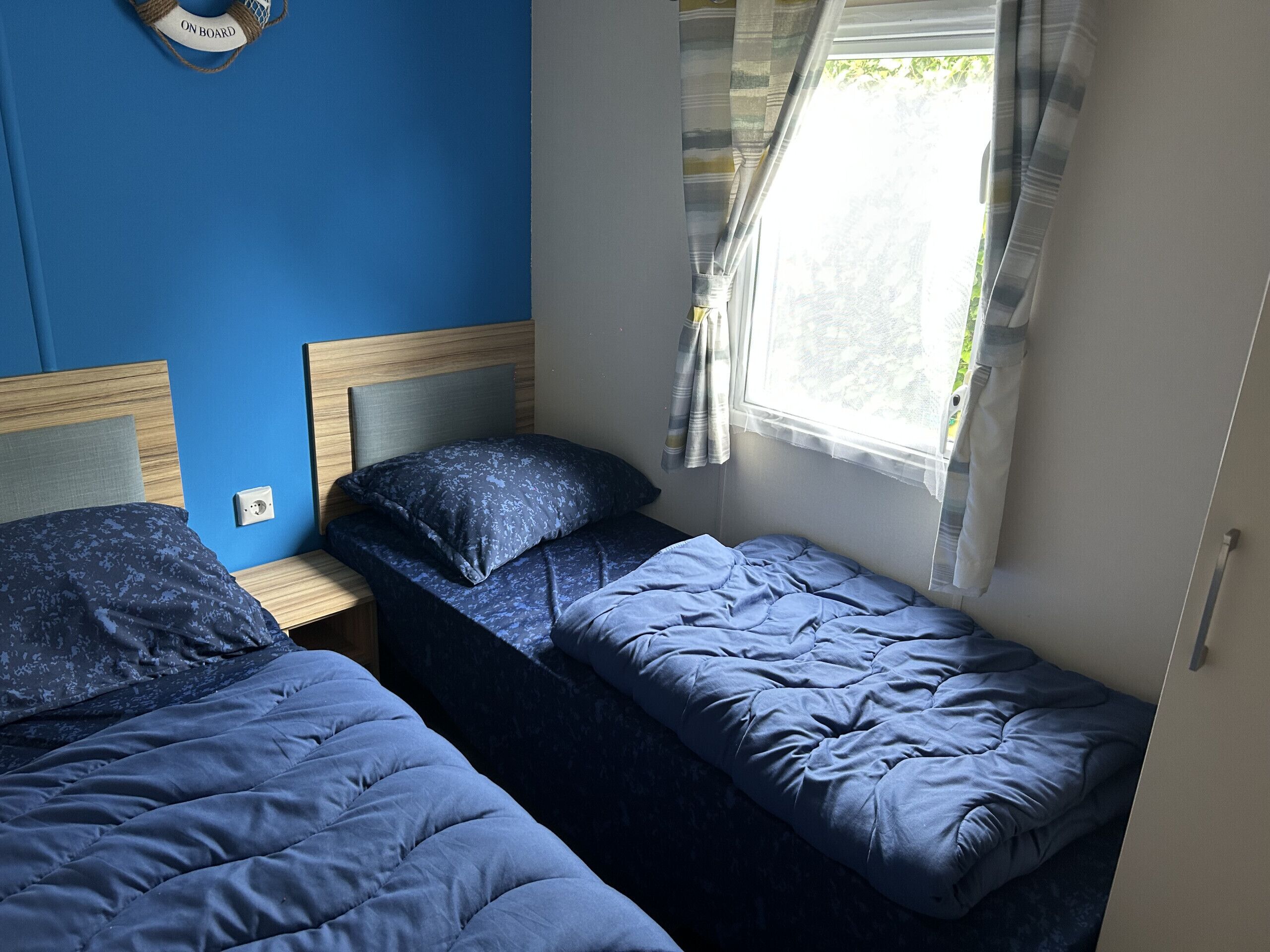 Una habitación con dos camas individuales, cabecera de madera, ventana con cortinas y un enchufe en la pared.