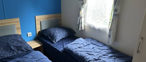 A room with two single beds, a wooden headboard, a window with curtains, and a wall socket.
