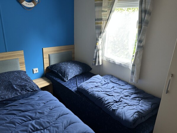 Una habitación con dos camas individuales, cabecera de madera, ventana con cortinas y un enchufe en la pared.