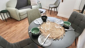 A dining area with a round table set for four, surrounded by modern chairs. A sofa and a chair are positioned against the wall, and a radiator is visible. - L'Olivier -4prs/ T2/Pontivy (Pontivy)
