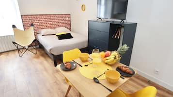 Une chambre d’hôtel moderne équipée d’un lit, d’une télévision et d’un coin repas avec une table dressée pour deux personnes, ainsi qu’un panier de fruits posé sur la table.