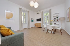 A modern living room with a grey sofa, a dining table with chairs, a flat-screen TV, and a bookshelf.